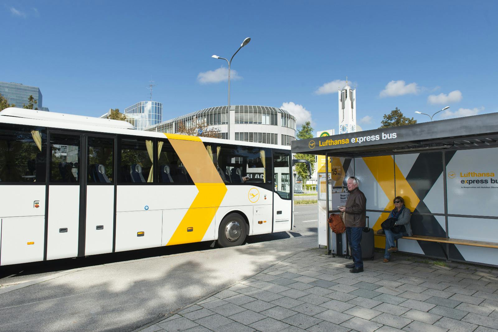 Two people wait at a Lufthansa Express bus stop; a white and yellow bus is parked in front. A modern building and clock tower are visible.