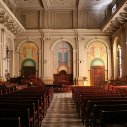 Interior of a church with wooden pews, high ceiling, and wall murals depicting a cross and religious symbols at the front.