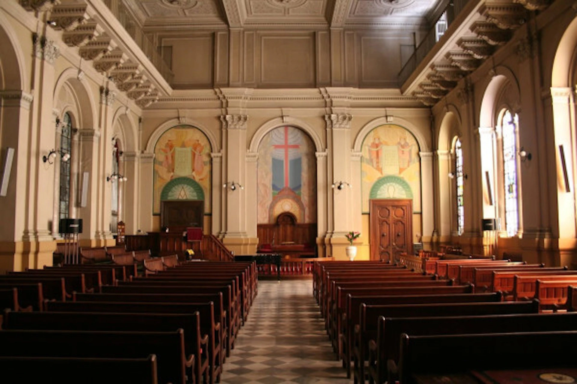 Interior of a church with wooden pews, high ceiling, and wall murals depicting a cross and religious symbols at the front.