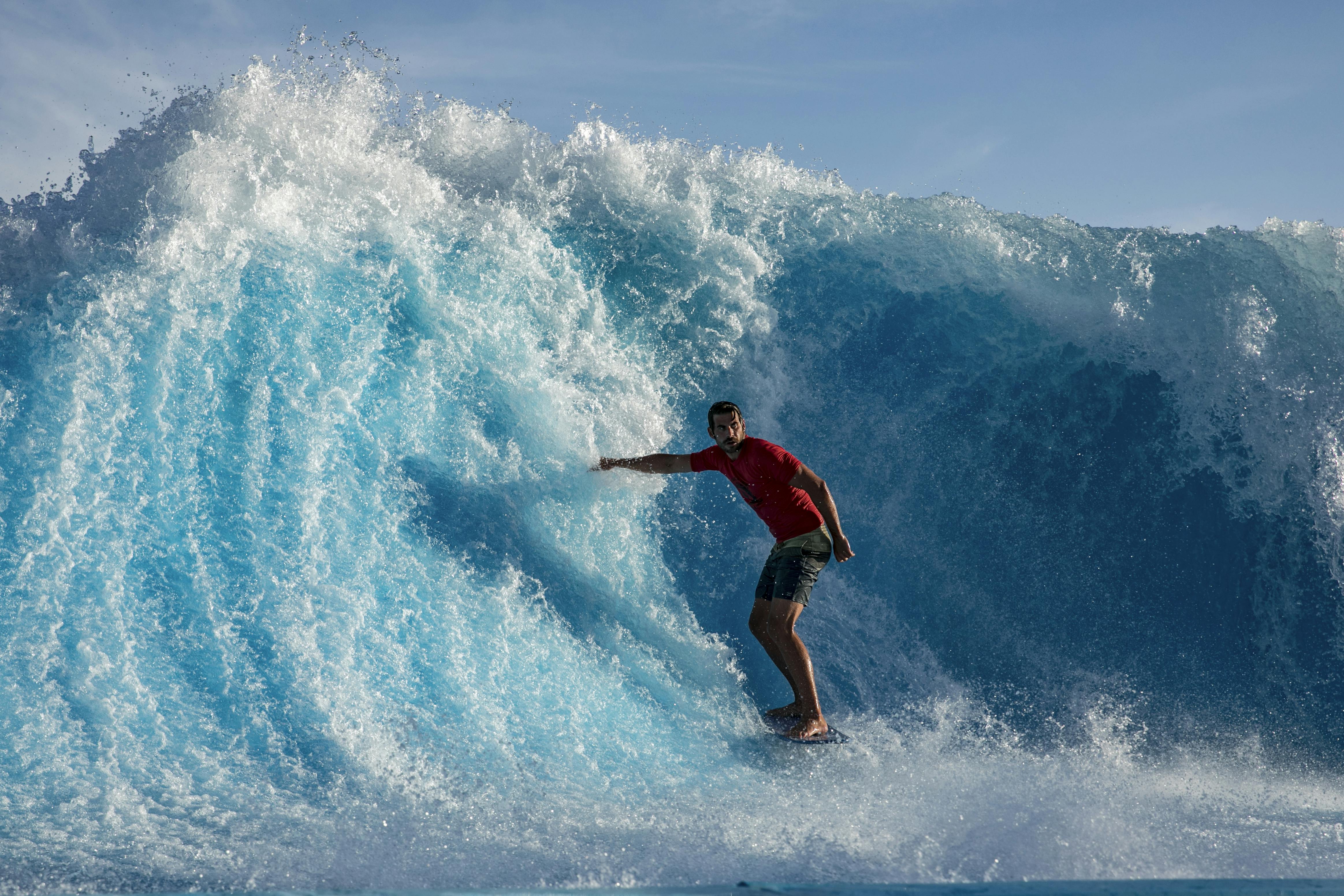 Person in a red shirt and shorts surfing on a large, blue ocean wave under a clear sky.