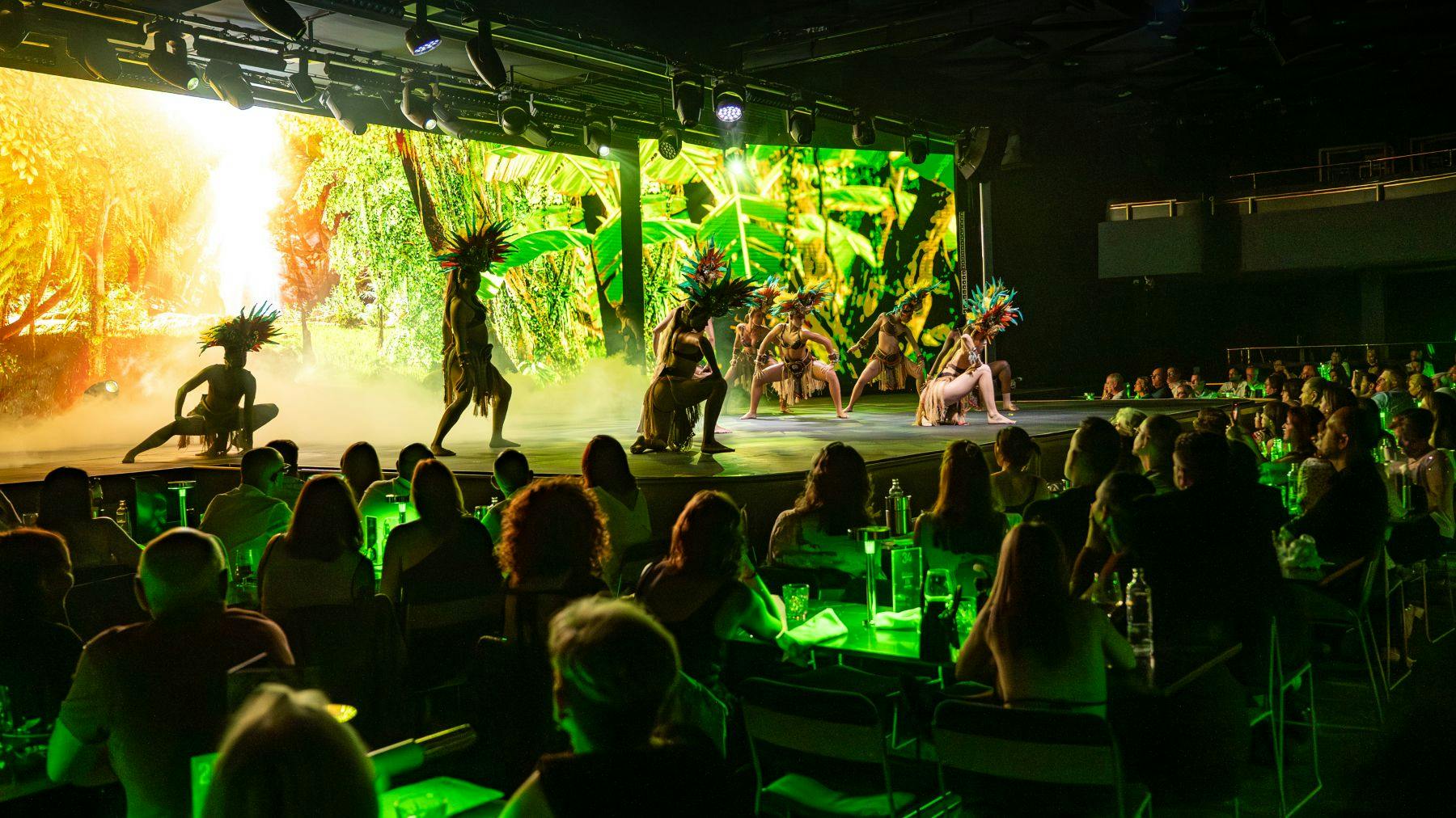 Artistas com elaborados toucados de penas dançam no palco com um cenário de selva, com o público sentado em primeiro plano.
