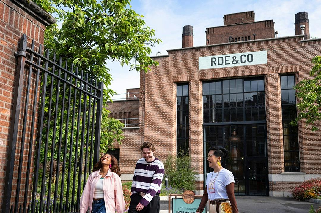 Three people smiling and walking outside a brick building with large windows and a sign reading "ROE & CO."