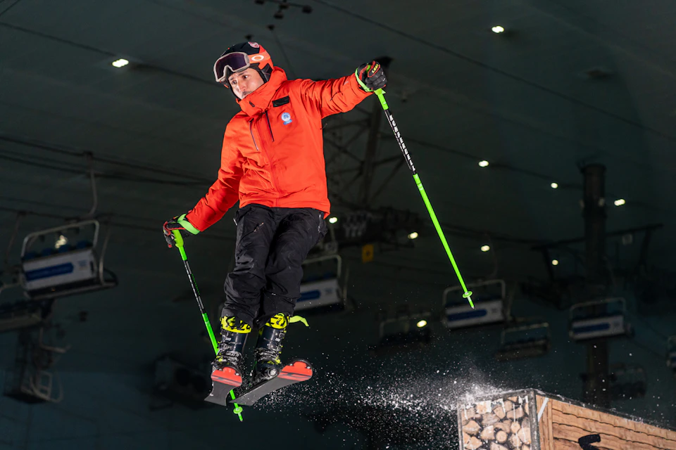 Person in an orange jacket and ski gear mid-air while skiing indoors, holding green ski poles.