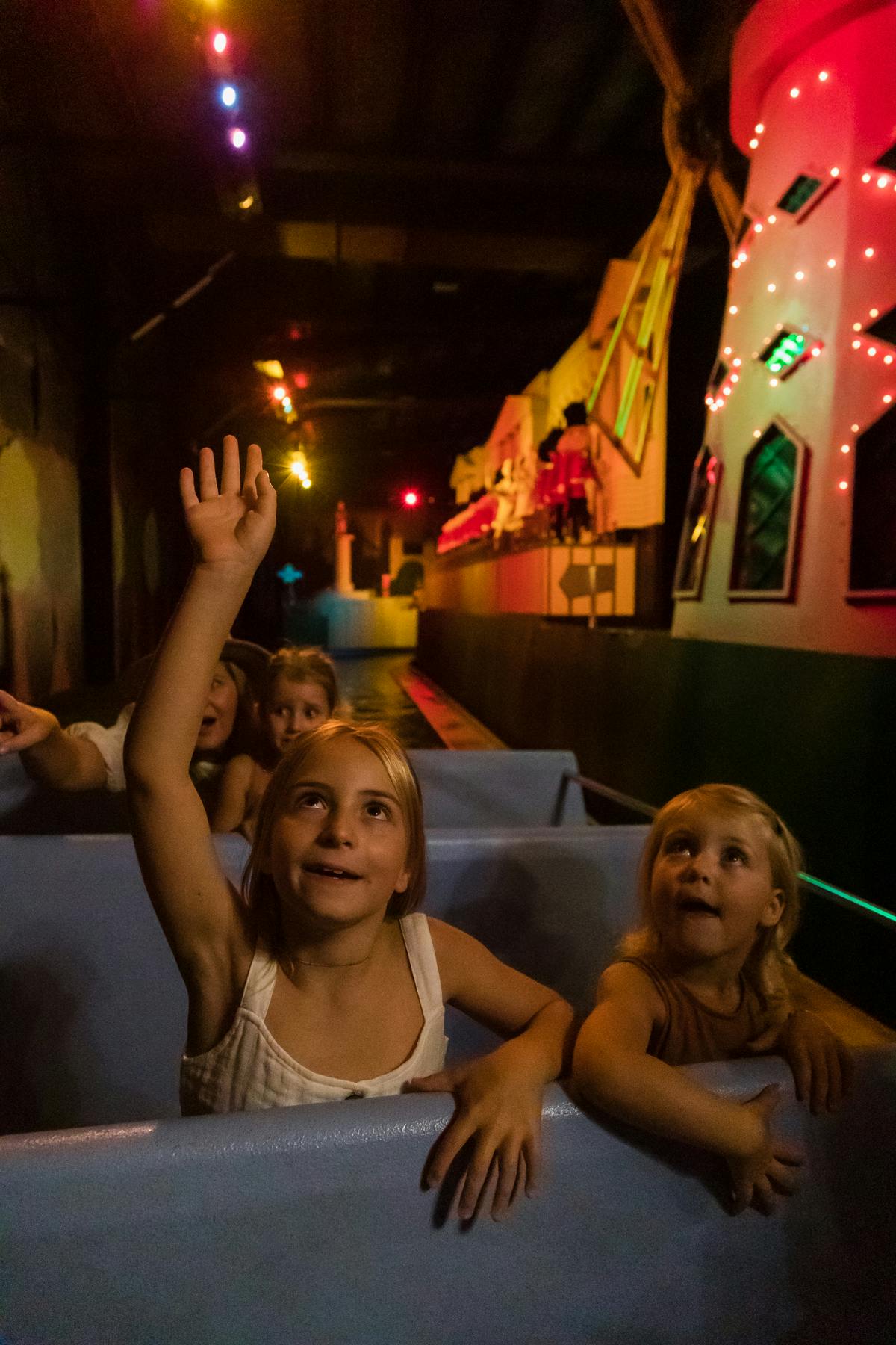 孩子们坐在黑暗的游乐设施中；一个女孩举手，另一些女孩兴奋地看着前方的动画场景。