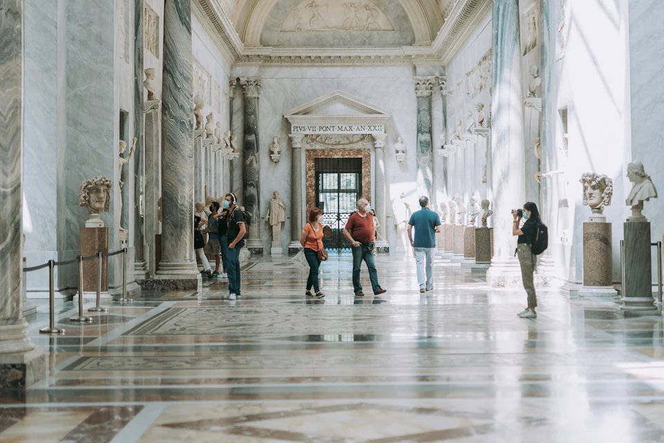 People walking and observing statues in a grand marble hall with high ceilings and ornate architectural details.