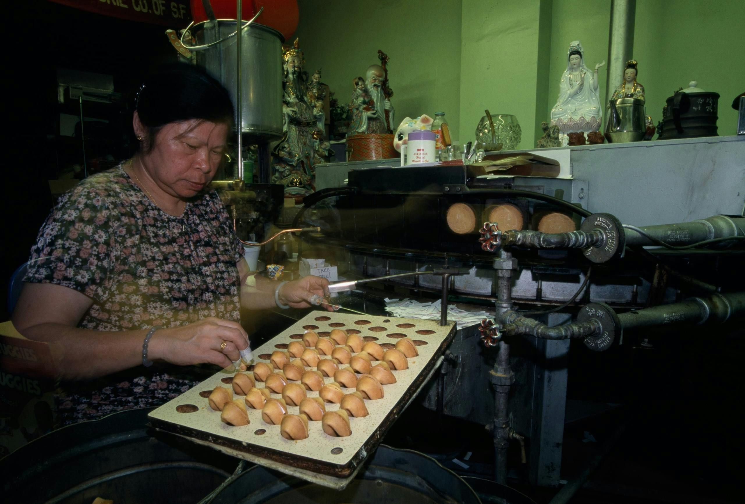 A woman preparing fortune cookies on a tray in a workshop with various equipment and decorations in the background.