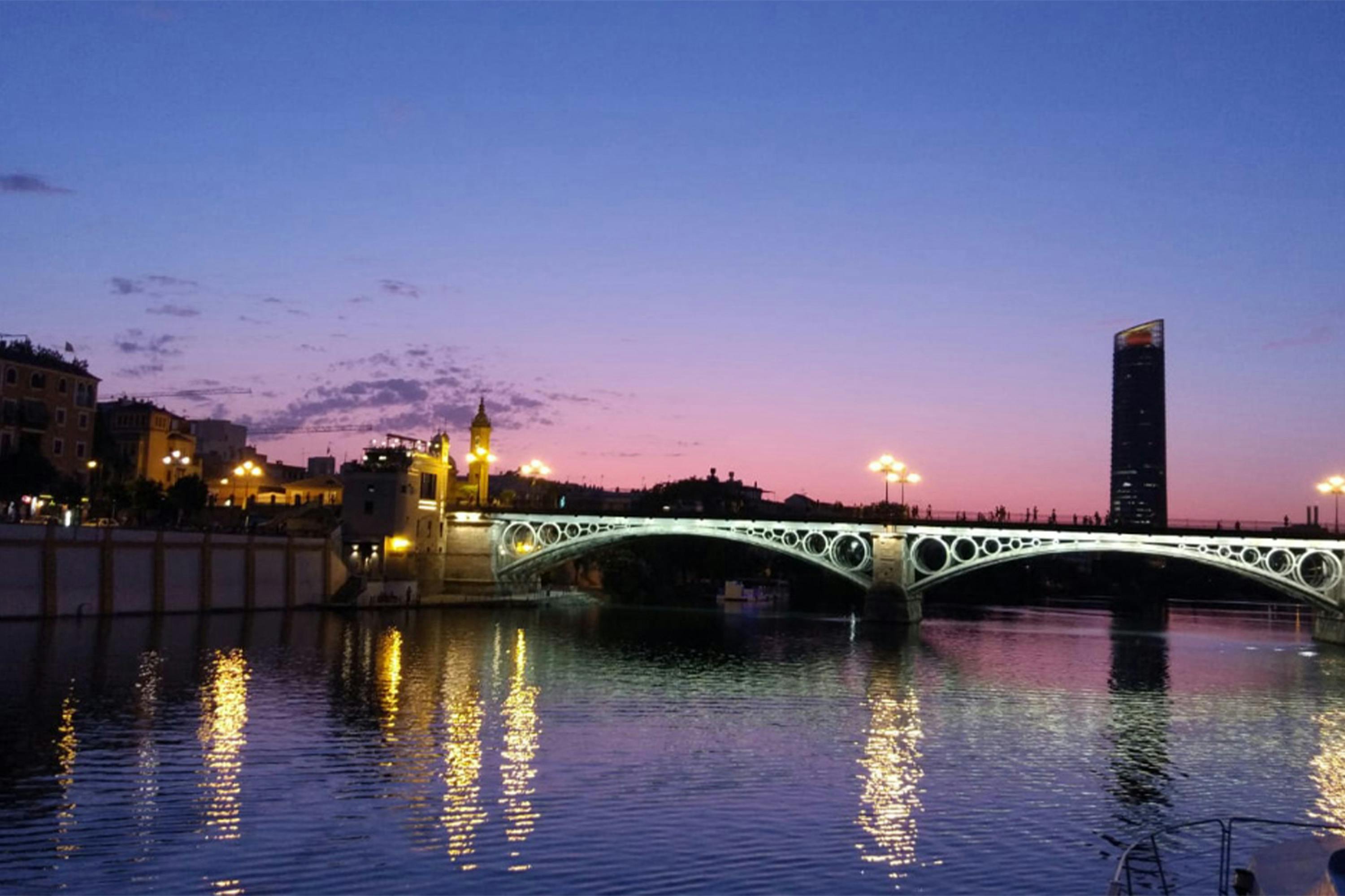 Podświetlany most z ozdobnymi łukami rozciąga się nad zbiornikiem wodnym o zmierzchu, z budynkami miejskimi i kolorowym zachodem słońca w tle.