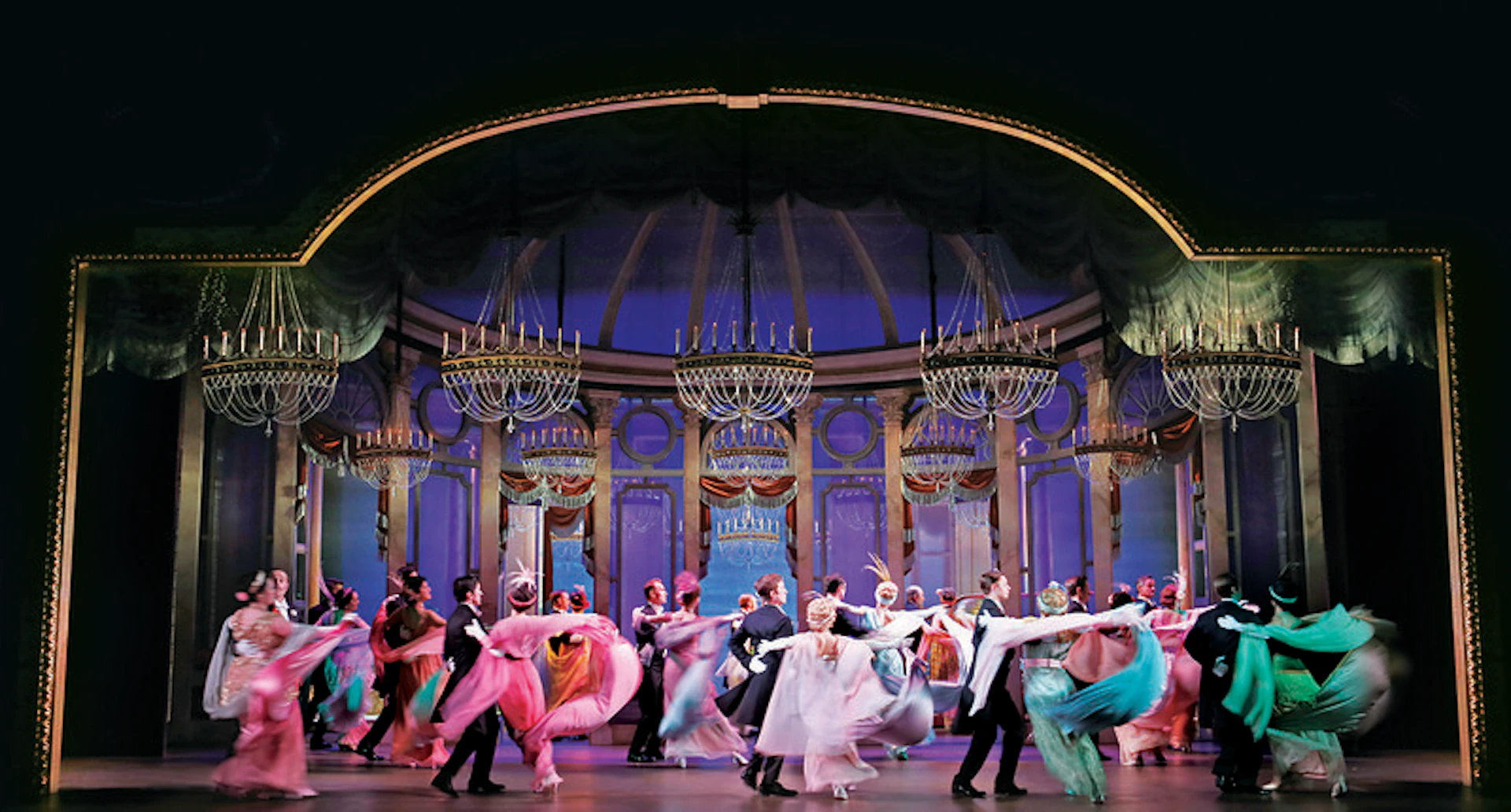 A group of performers in elegant attire dancing on a theater stage with an ornate backdrop and chandeliers.