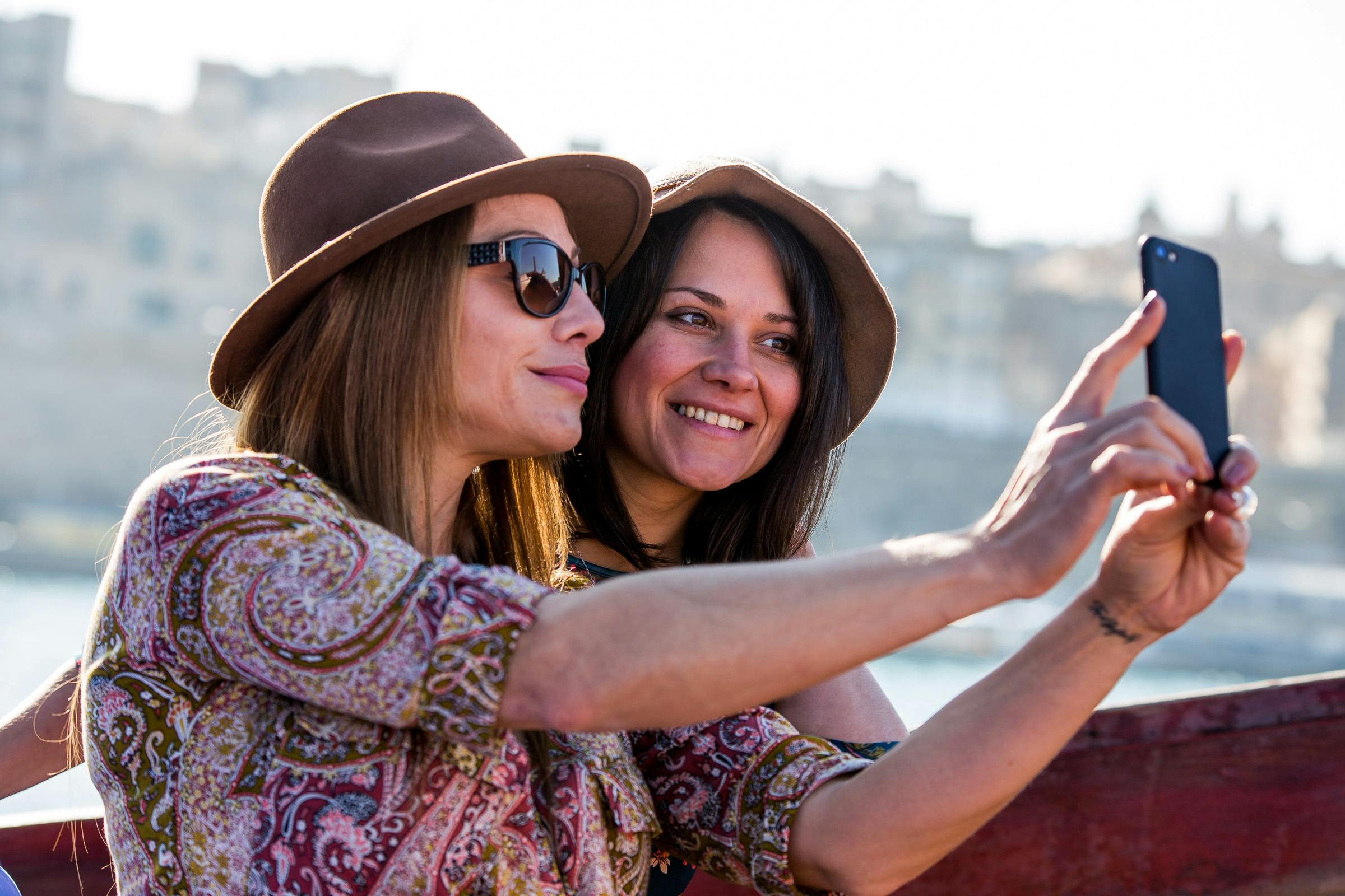 ぼやけた背景の屋外で自撮りをしながら、帽子をかぶり、模様入りの服を着た2人の女性が微笑んでいます。