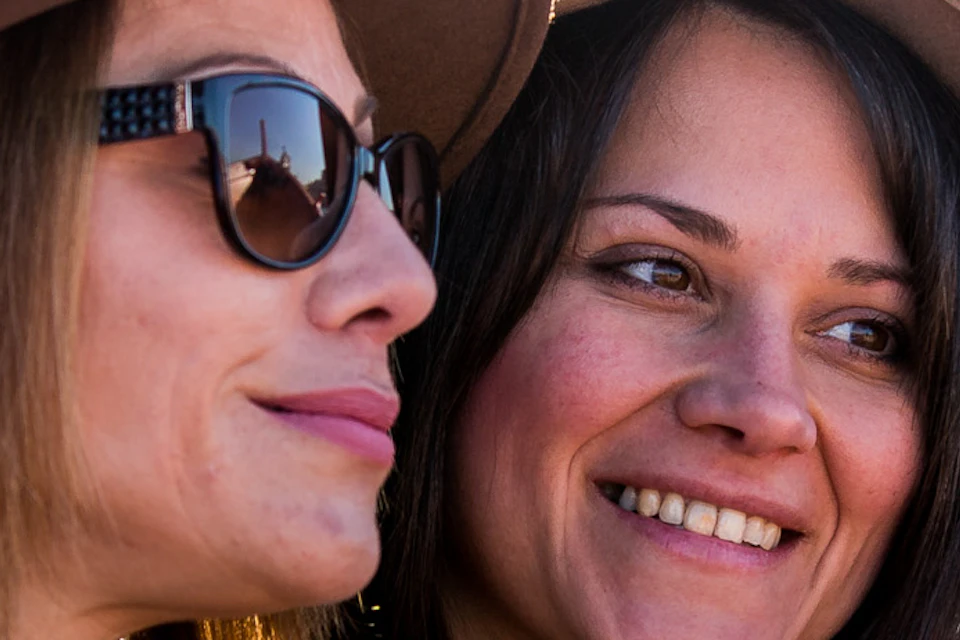 Two women wearing hats and patterned clothing smile while taking a selfie outdoors with a blurred background.