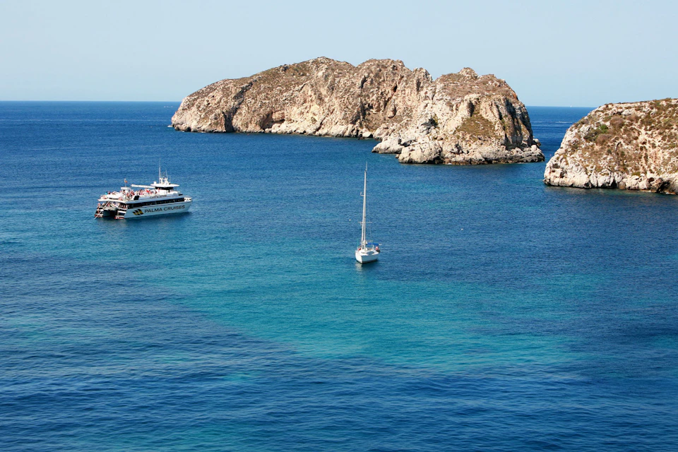 A sailboat and a larger boat near a rocky island in a calm, blue sea under a clear sky.
