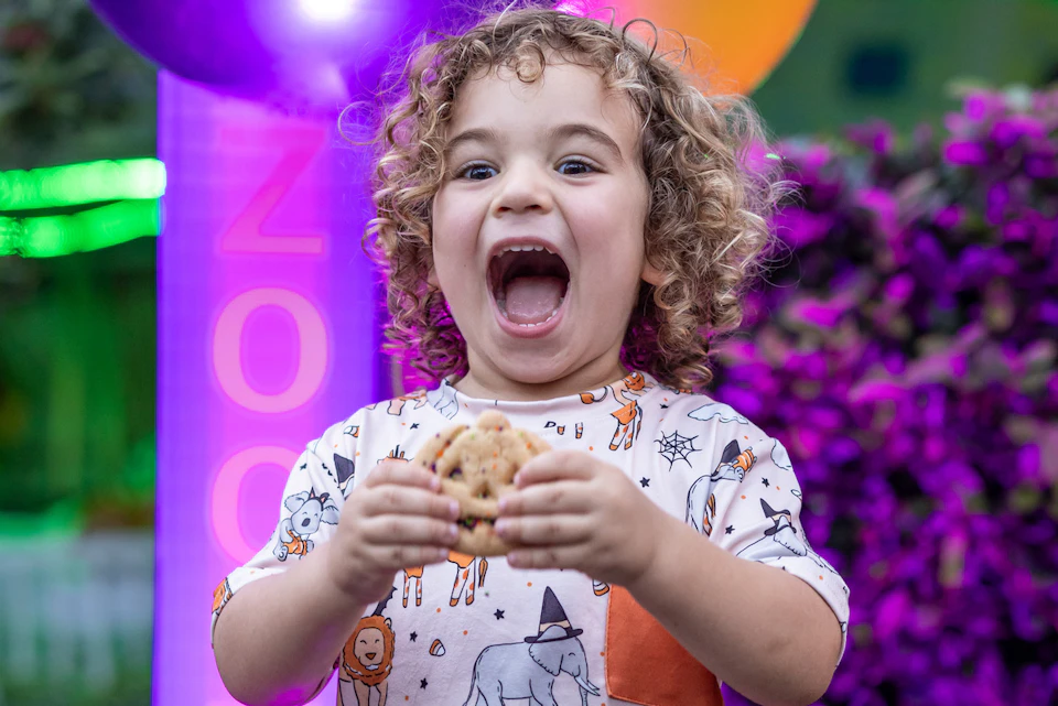 A young child with curly hair is excitedly holding a cookie, wearing an animal-themed shirt in a vibrant, colorful setting.