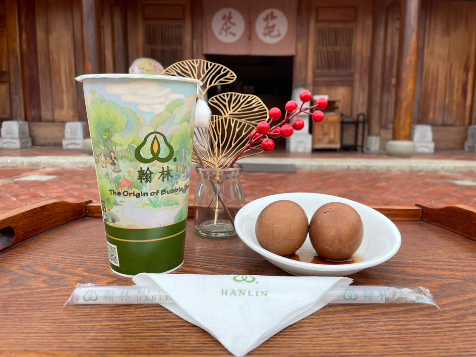 A cup with bubble tea branding, a bowl with two tea eggs, and some decorative items on a table in front of a wooden building.