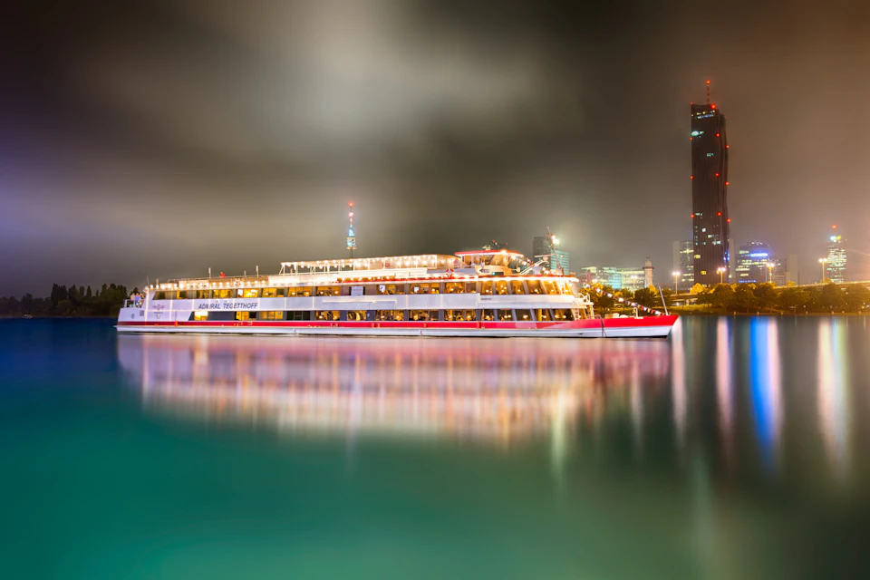 A brightly lit cruise ship gliding on calm water at night, with cityscape and tall buildings illuminated in the background.