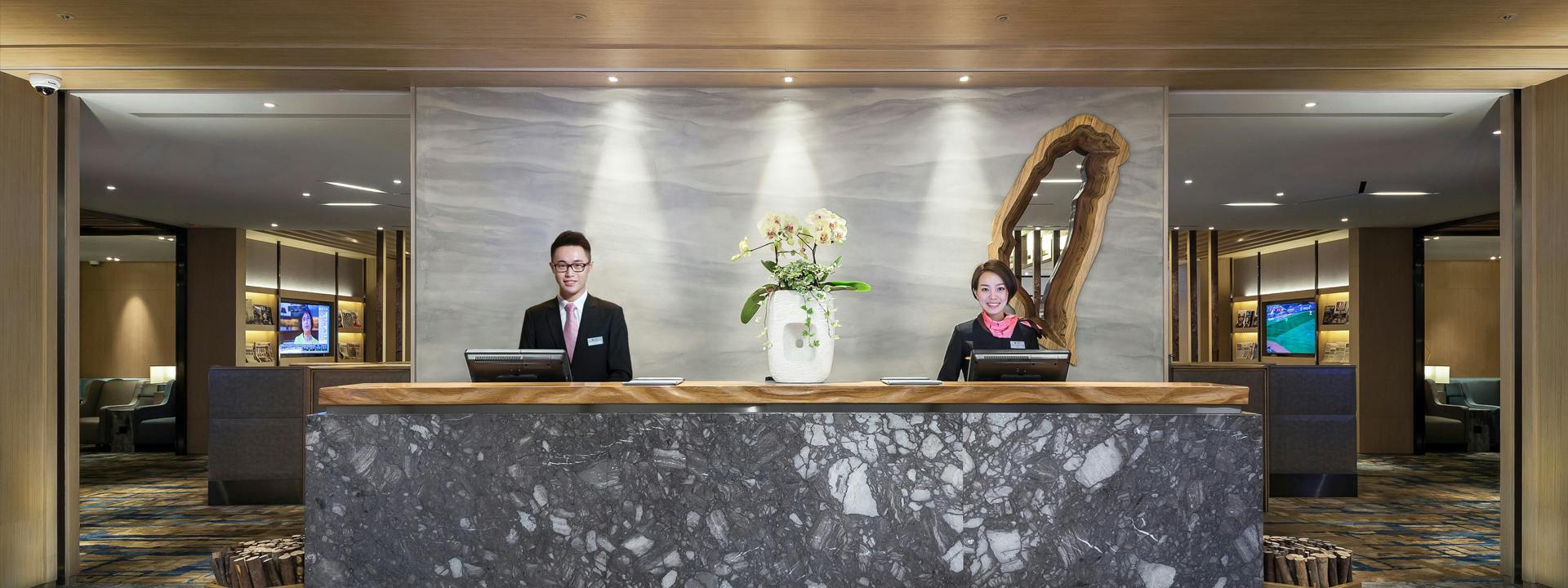 Recepcja hotelowa z dwoma pracownikami stojącymi za nią, elementami dekoracyjnymi i częścią wypoczynkową w tle.