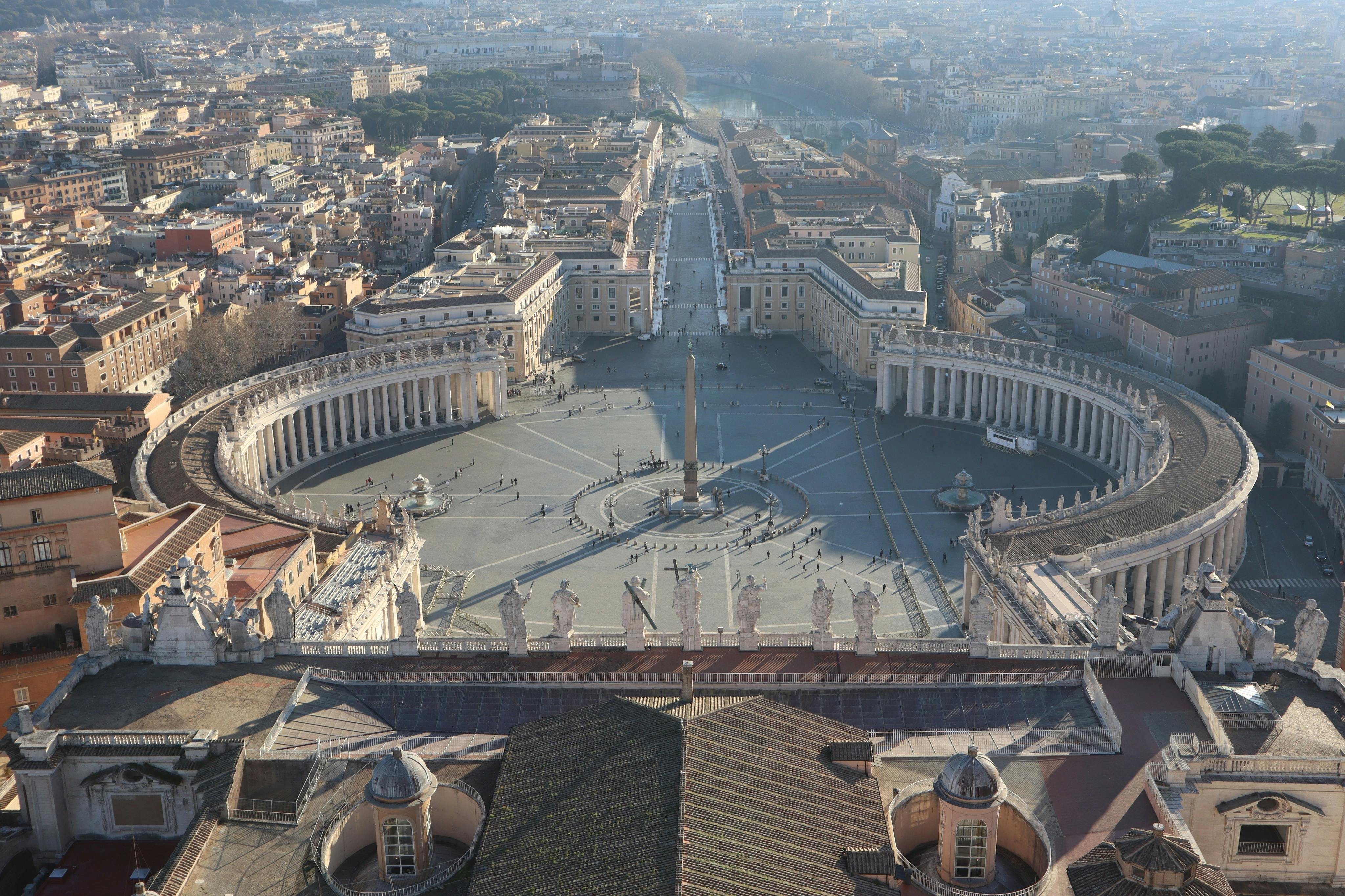 Veduta aerea di una grande piazza con un obelisco centrale, circondato da colonnati curvi e un paesaggio urbano sullo sfondo.