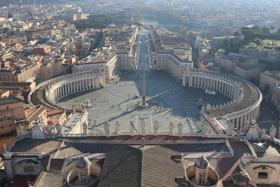 Aerial view of a large plaza with a central obelisk, surrounded by curved colonnades, and a cityscape in the background.
