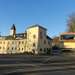 En historisk byggnad med ett kyrktorn bredvid Alpenzoo-ingången under en klarblå himmel, med en tom asfalterad yta i förgrunden.