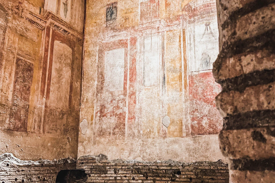 A weathered wall with faded frescoes depicting architectural elements and figures, located in an ancient brick structure.