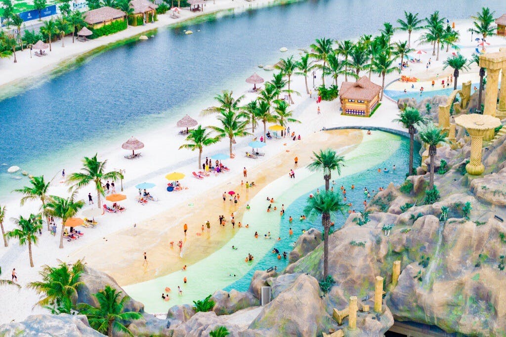 Aerial view of a tropical water park with a sandy beach, people swimming, lounging under umbrellas, palm trees, and rocky structures.