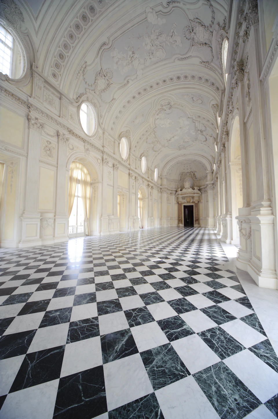 An ornate, long hall with a checkered marble floor, arched windows, and detailed vaulted ceiling with intricate designs.