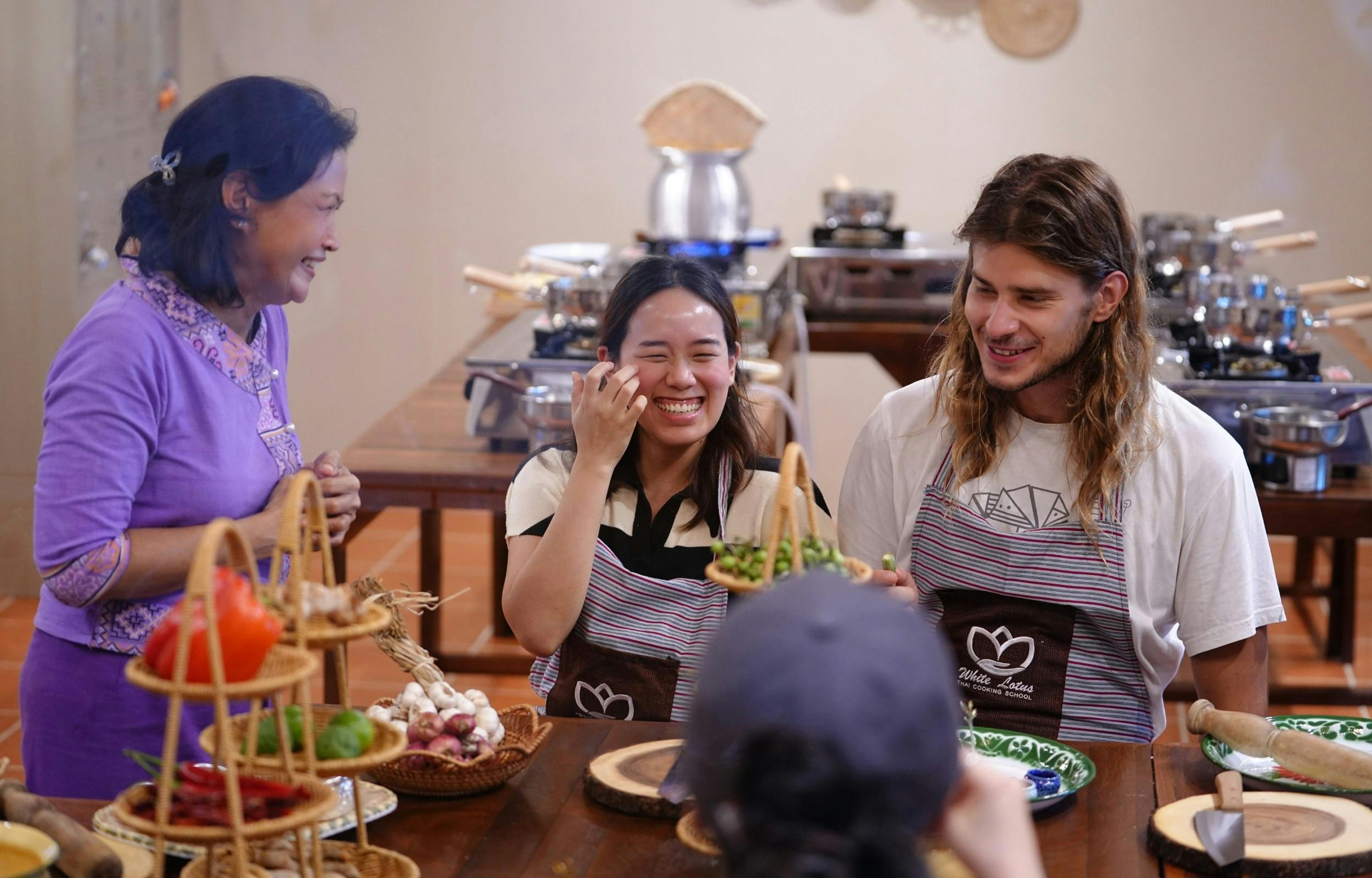 A group of people in a cooking class laughing and interacting around a table with baskets of vegetables and cooking materials.
