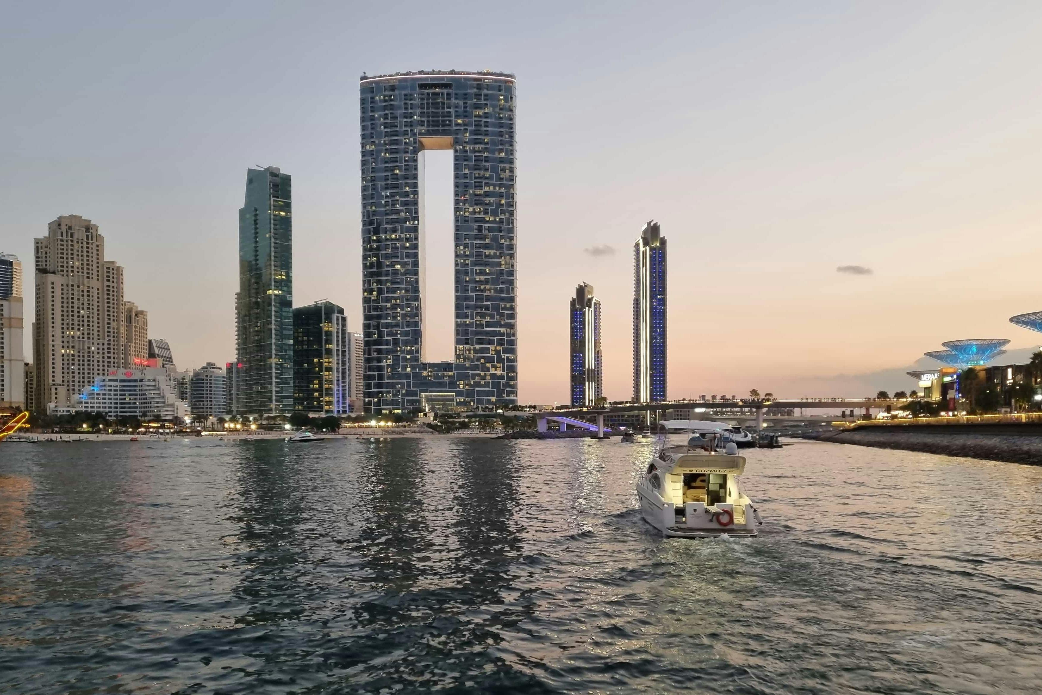 A motorboat on water near a cityscape with tall, lit-up modern buildings at sunset.