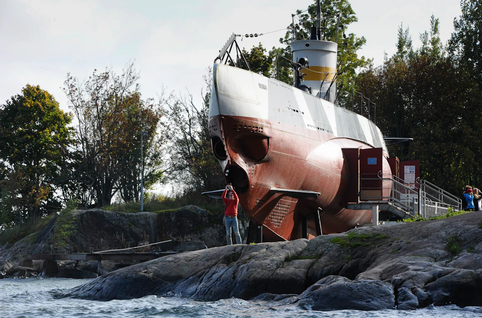 A man in a red shirt stands on rocky shore, photographing a large submarine displayed on land with trees in the background.
