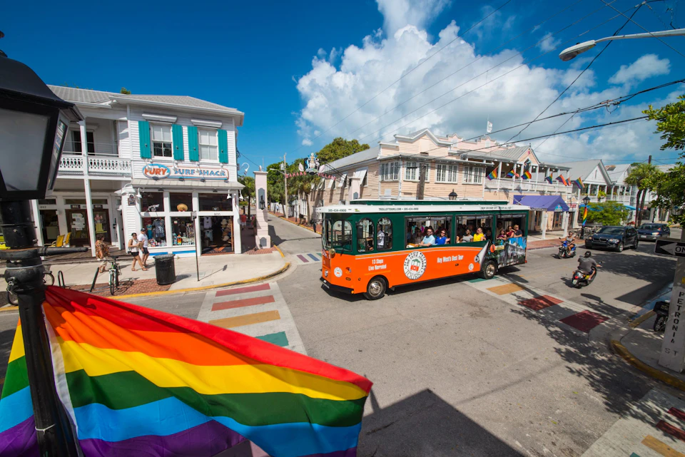 A street scene with a green and orange tour trolley, a rainbow flag, and colorful buildings under a bright blue sky.