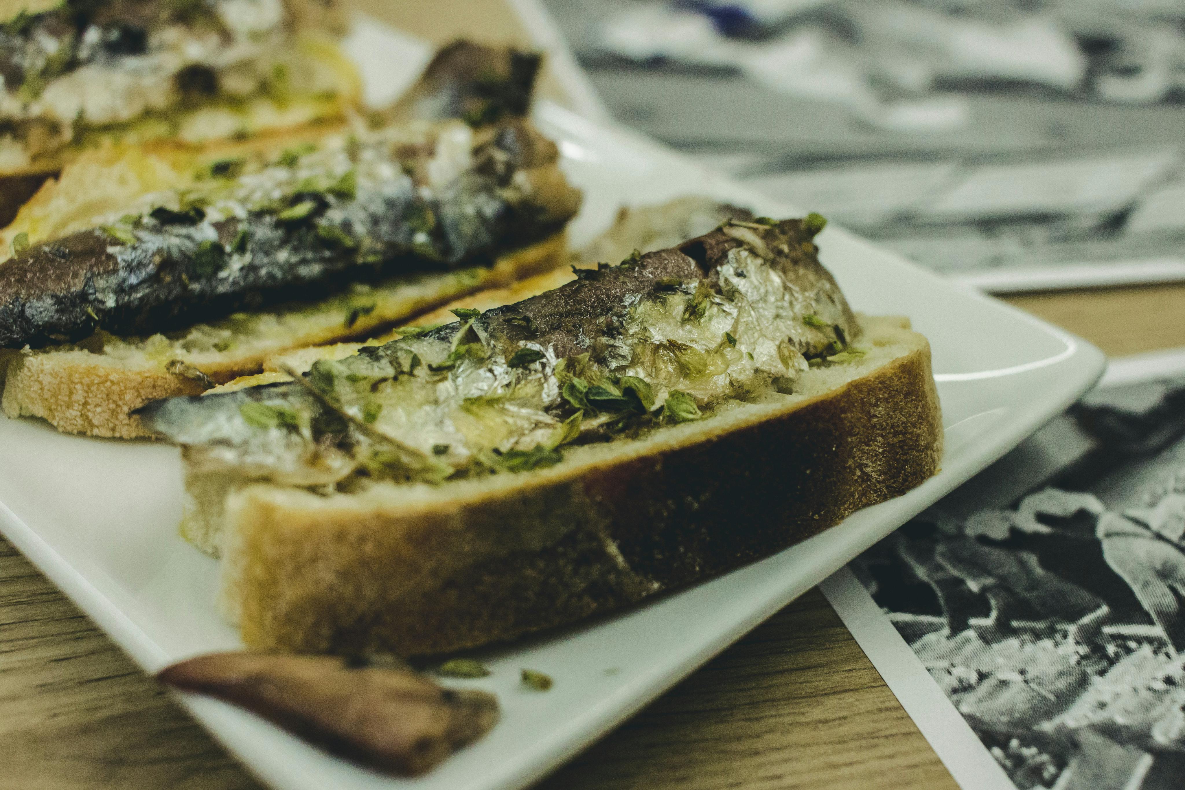 Plakjes brood belegd met gegrilde sardientjes en kruiden op een wit bord op een houten tafel.