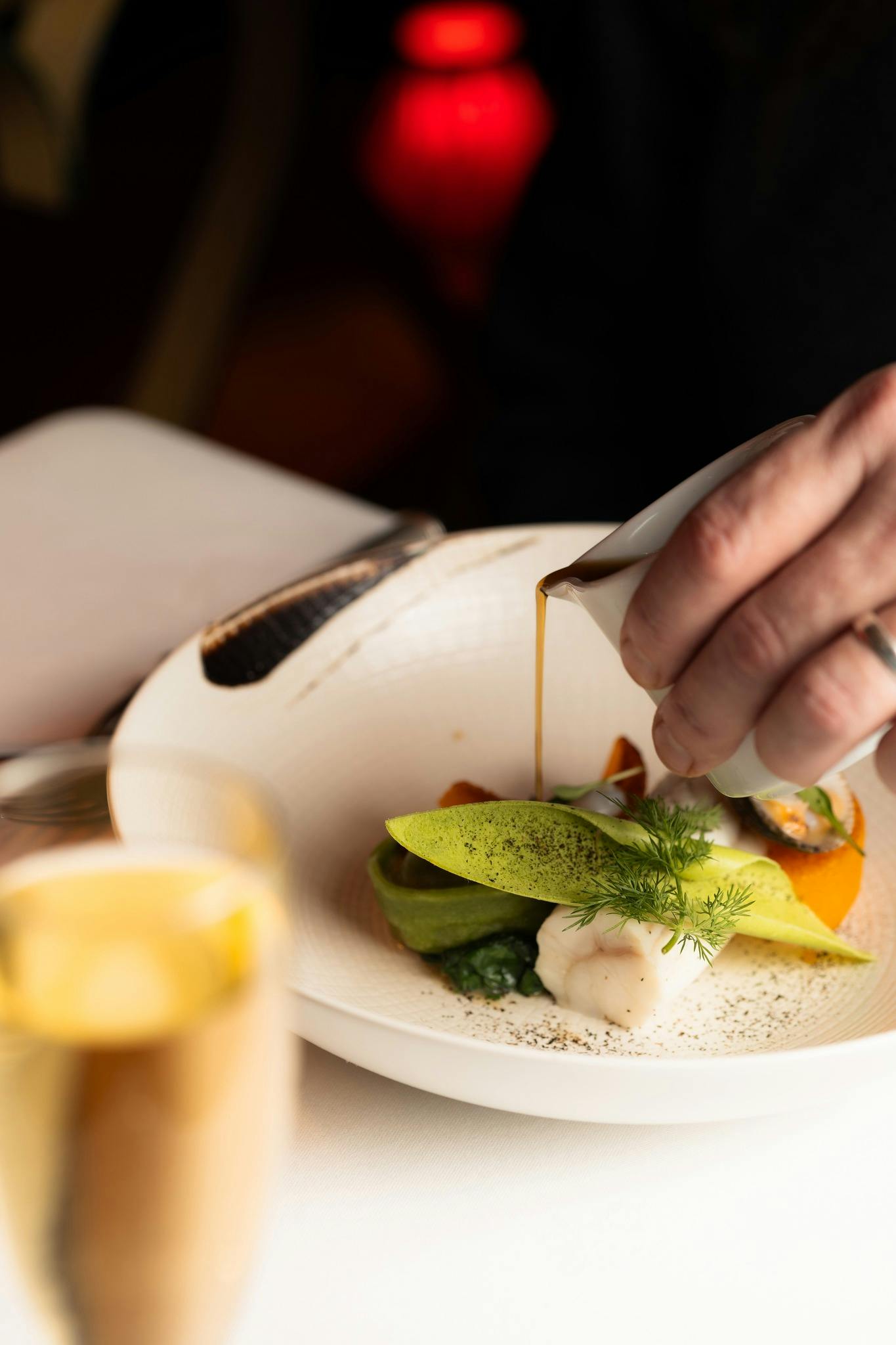 A person pouring sauce onto a gourmet dish with fish, vegetables, and a garnish on a white plate.