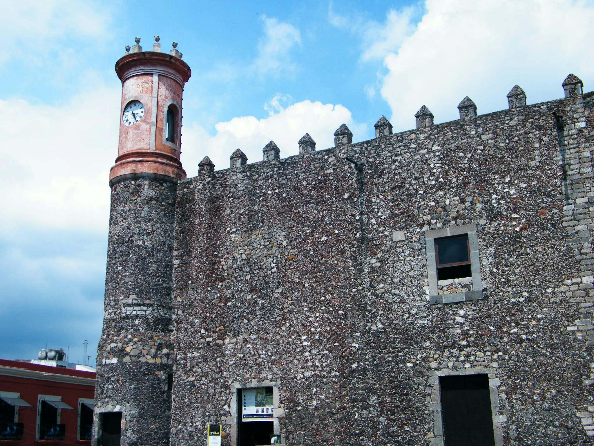 Kamienny budynek z wieżą zegarową, krenelażowym dachem i oknem pod błękitnym niebem. Częściowo widoczny jest kolejny budynek i znaki.