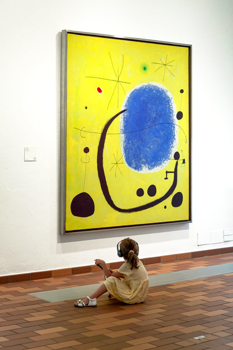 A child in a yellow dress sits on the floor, wearing headphones and looking up at a large abstract painting with yellow background and blue and black shapes.