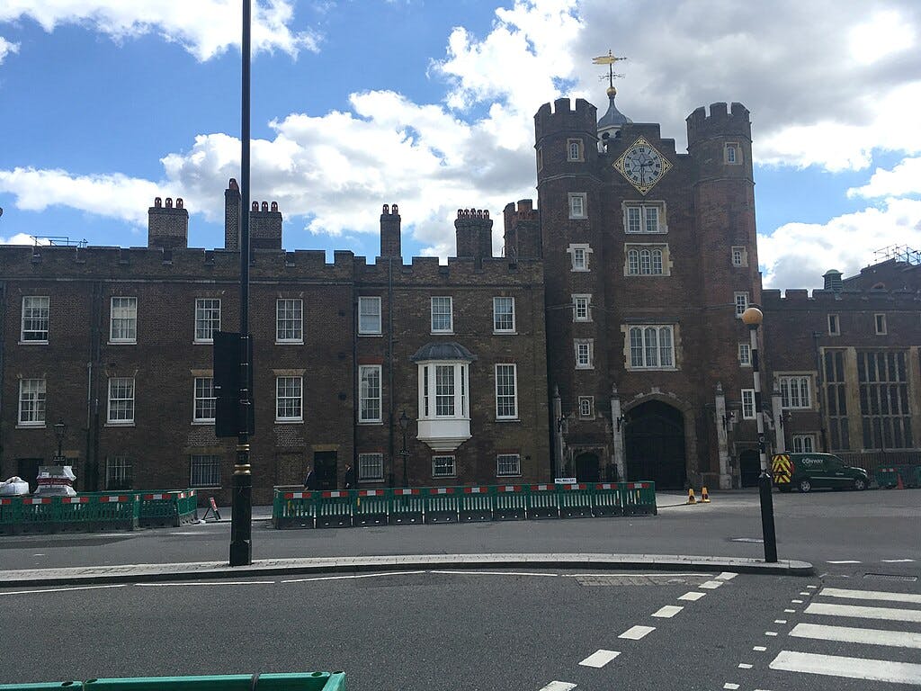 Duży murowany budynek z centralnym łukowym wejściem, zegarem i wieżami, otoczony barykadami i ulicą na pierwszym planie.