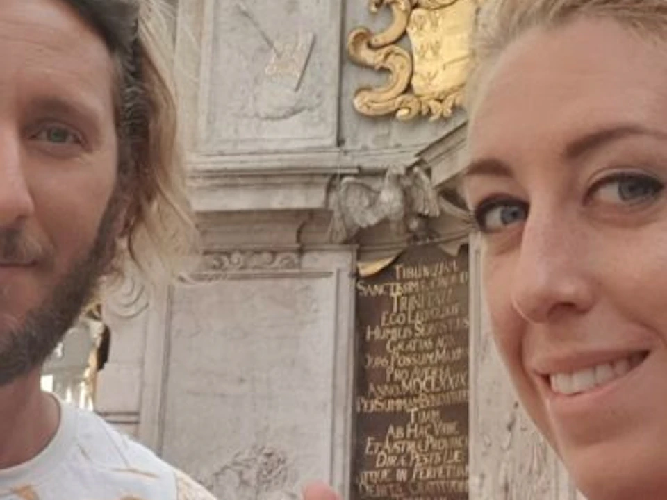 A man and woman pose for a selfie in front of a monument with intricate carvings and plaques. The man gives a thumbs-up gesture.