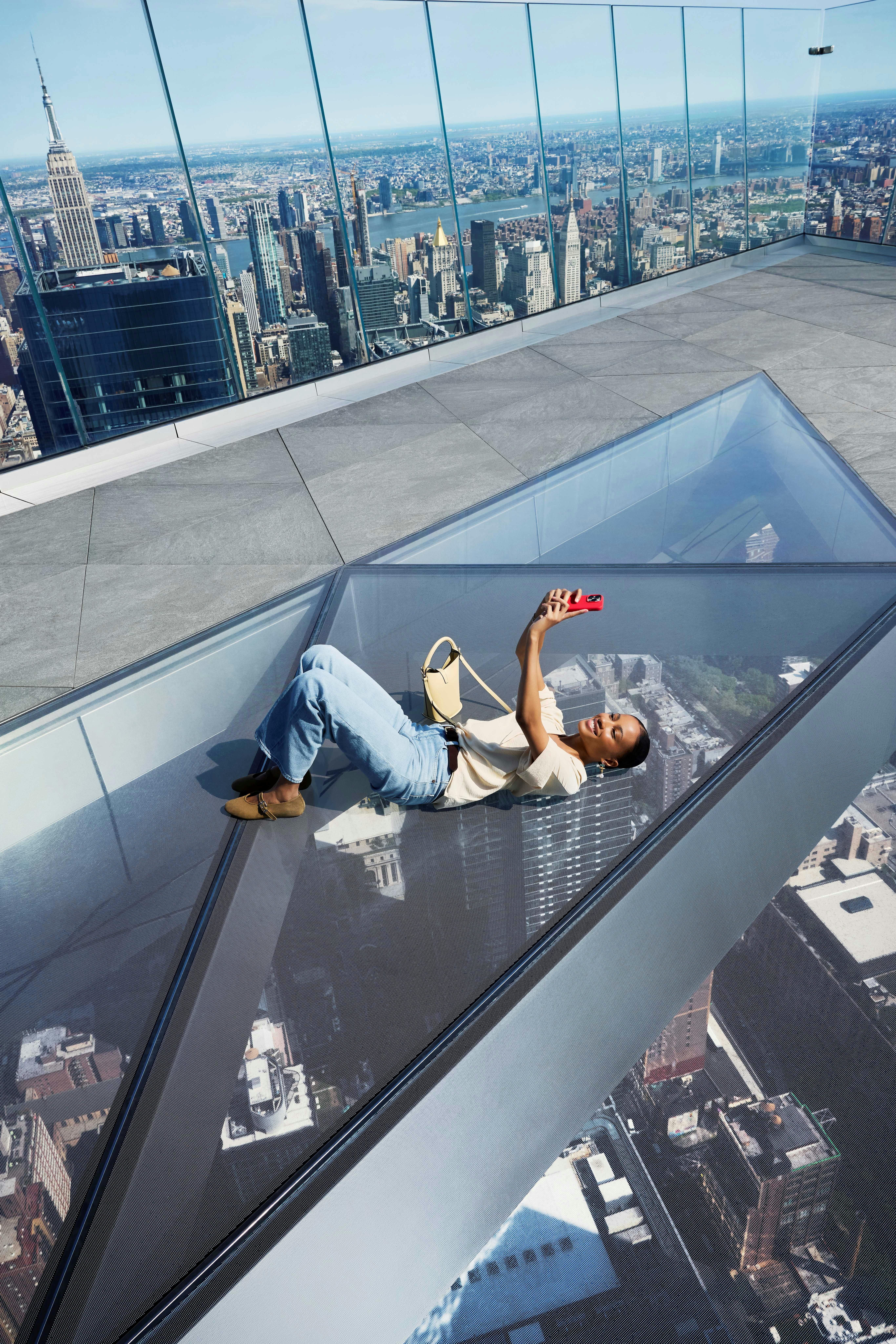 Person lying on a glass floor of a tall building, taking a selfie with a cityscape visible below.