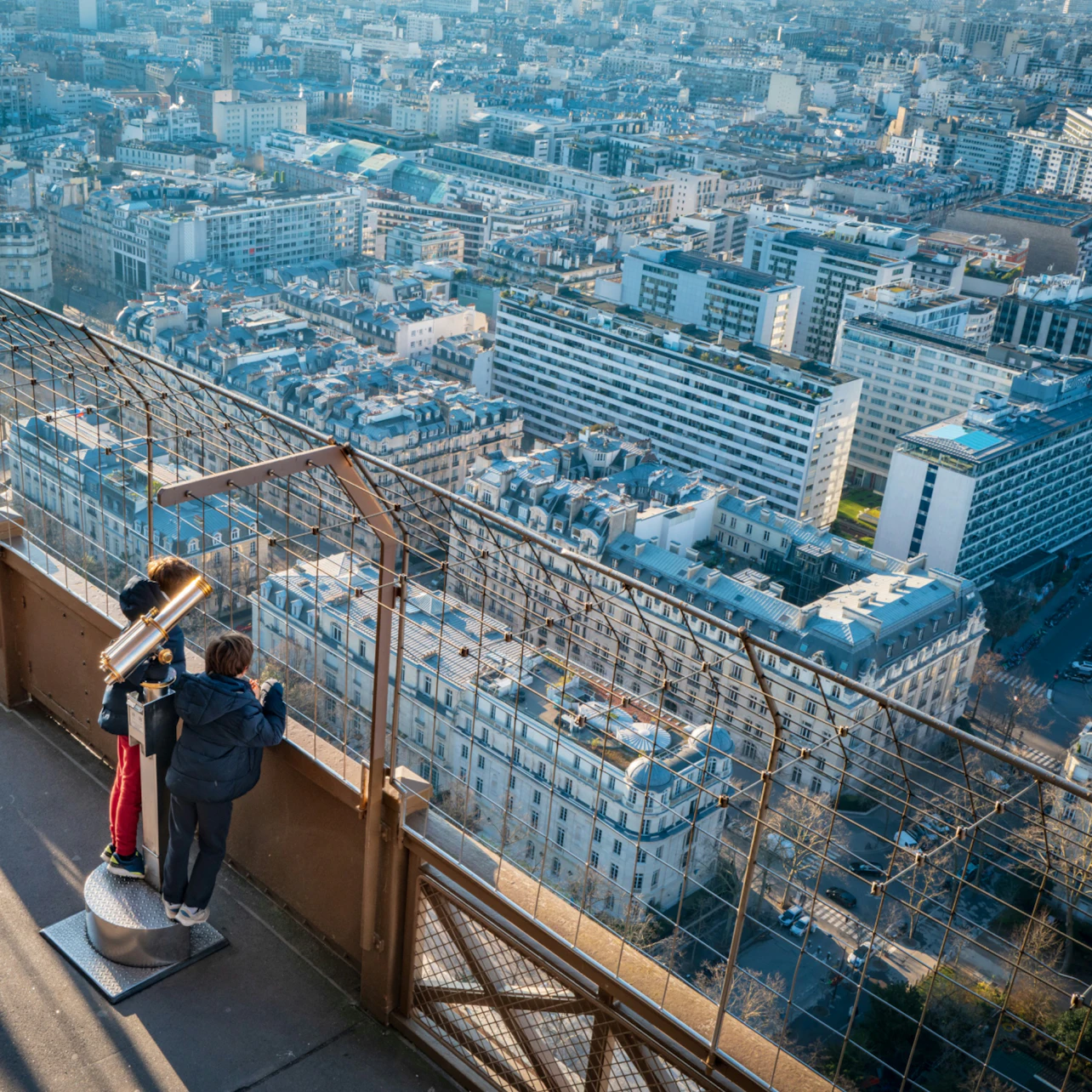 Two people on an observation deck looking at a cityscape through a telescope, with a metal fence in the foreground.