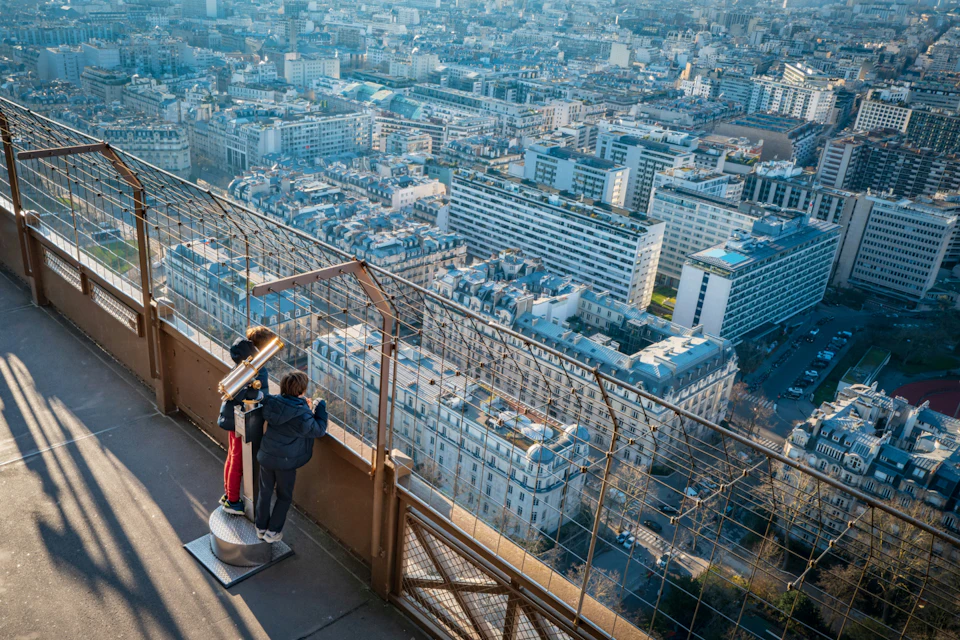 Two people on an observation deck looking at a cityscape through a telescope, with a metal fence in the foreground.