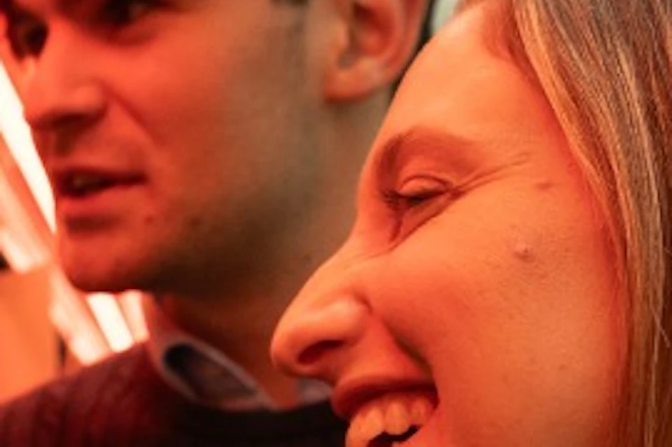 Two people smiling and interacting at a counter in a warmly lit indoor setting with glass and wood decor.