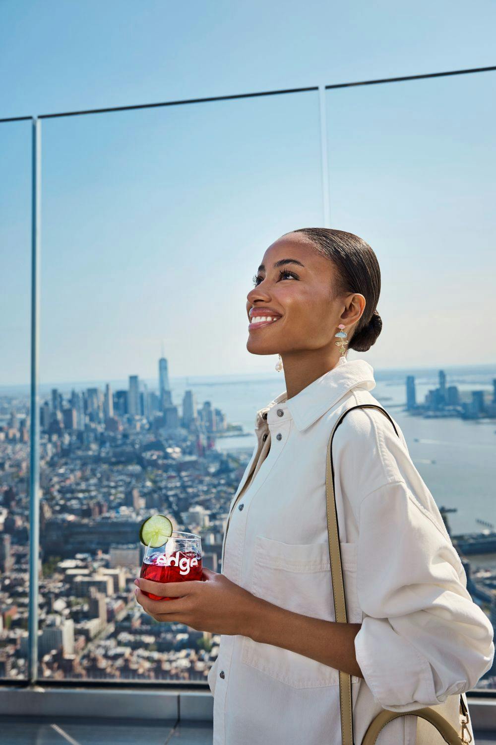 A person smiling in profile, standing on a rooftop with a view of a city skyline and a sunny, clear sky in the background.