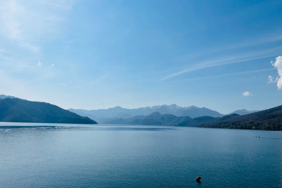 A serene lake with calm water surrounded by distant mountain ranges under a clear blue sky.