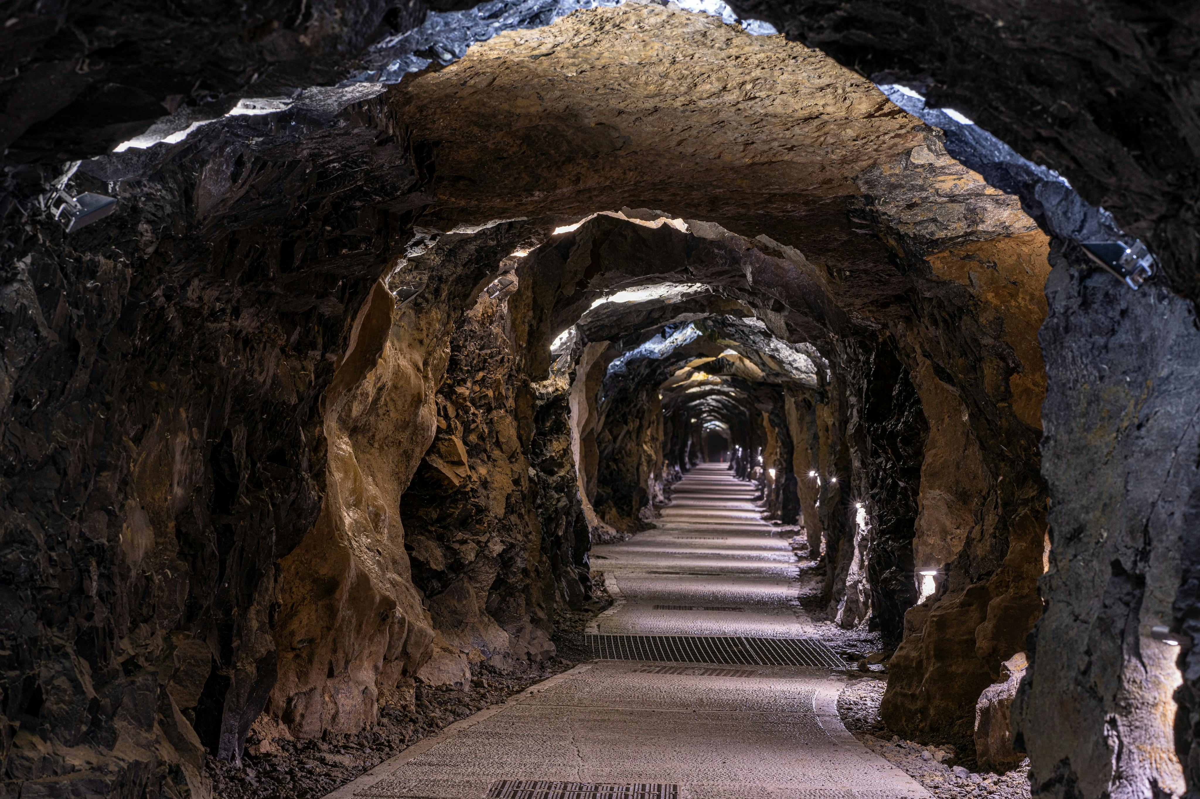 A rocky underground tunnel with a lit pathway extending into the distance, featuring rough stone walls and an arched ceiling.