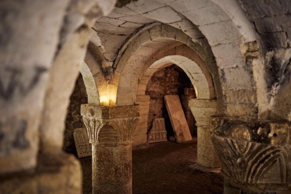Ancient stone arches and columns in a dimly lit underground room, with scattered stone slabs and textured brick walls.