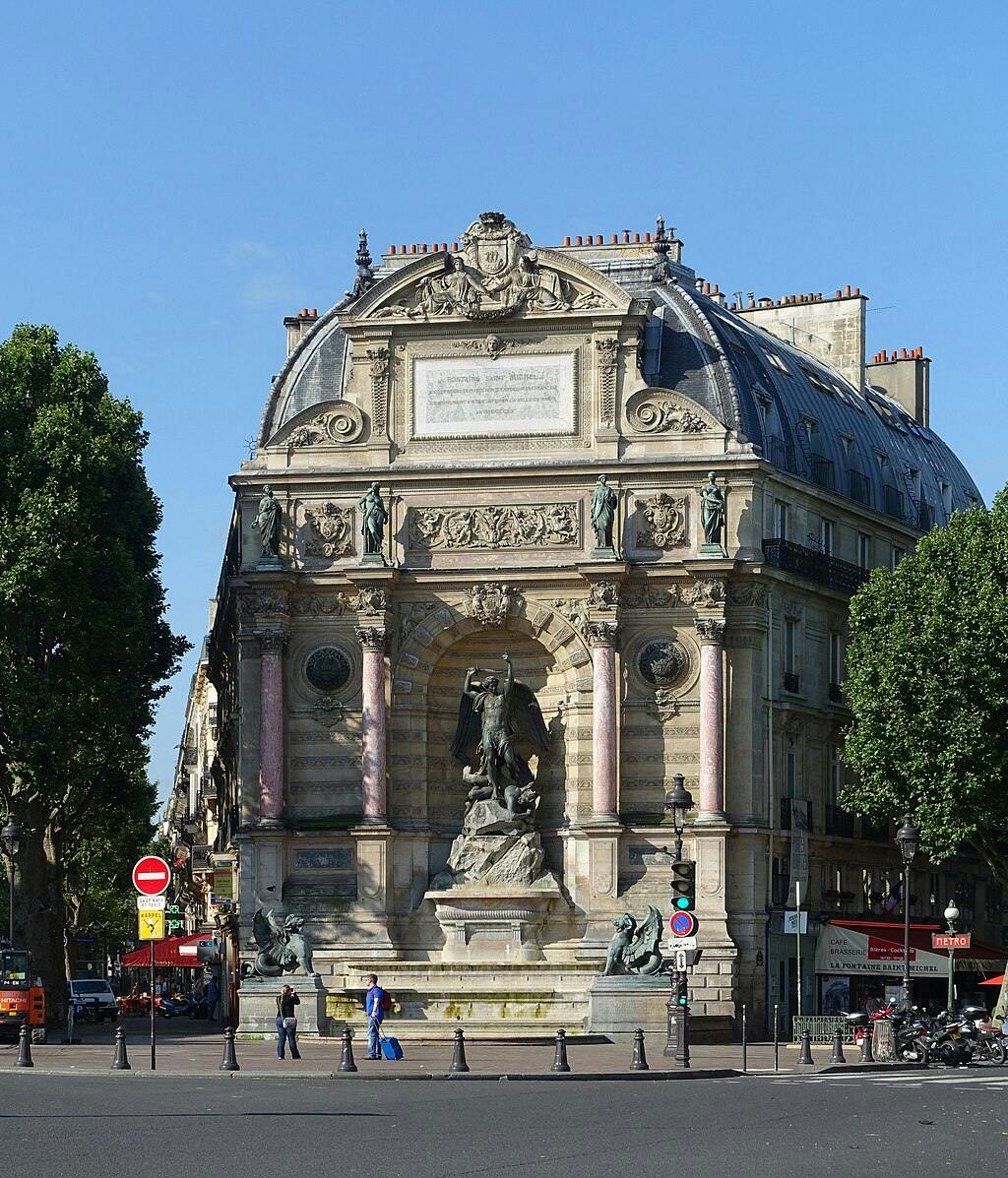 Fontaine historique et ornée de statues, grand bâtiment derrière, arbres sur les côtés, personnes marchant, ciel dégagé.