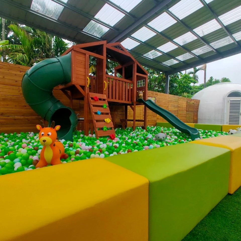 Children's play area with a wooden playset, slides, and a ball pit, under a roofed structure surrounded by greenery.