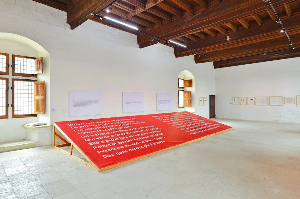 Large red platform with white text in French, set in a spacious room with white walls, artworks, and wooden ceiling beams.