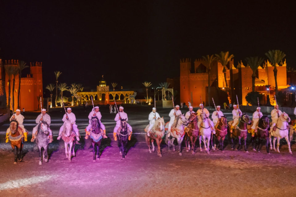 A group of people on horseback, dressed in traditional attire, lined up in front of illuminated buildings and palm trees at night.