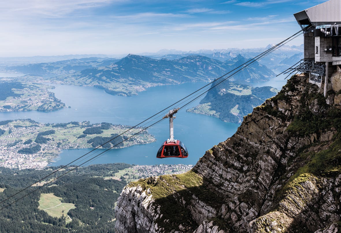 A red cable car travels over a steep mountain cliff with a scenic view of a lake and distant green hills below.