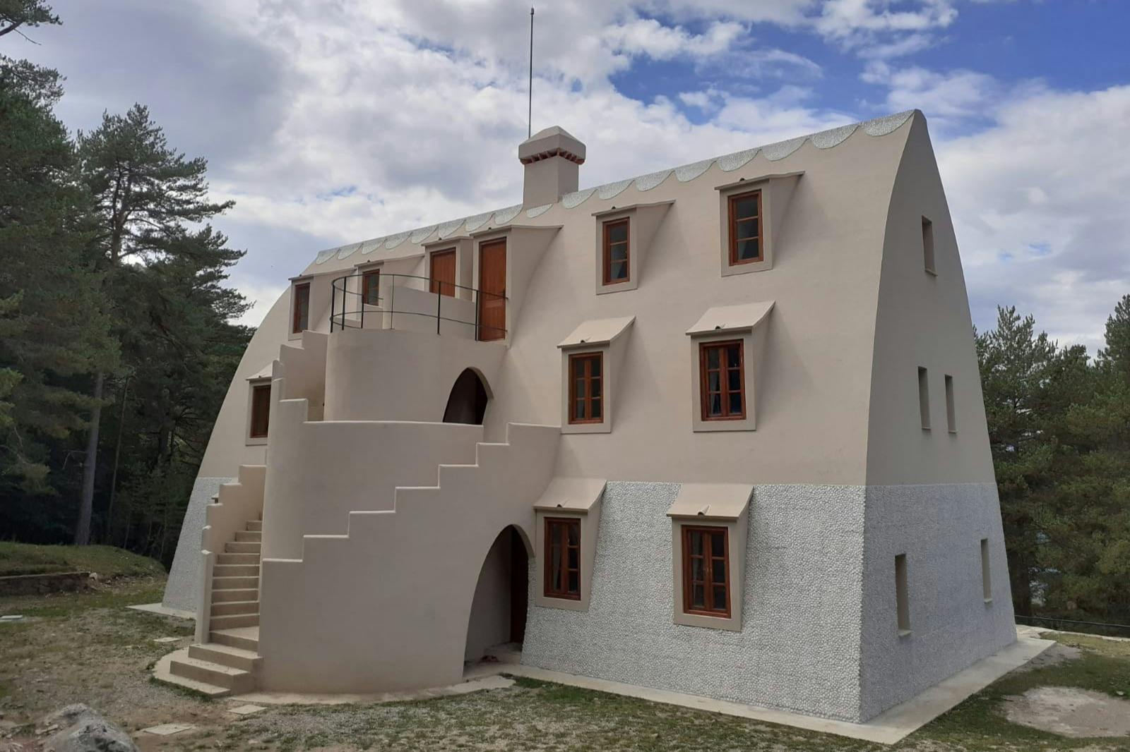 Un bâtiment beige unique en son genre, doté d'un toit incurvé, de nombreuses fenêtres et d'un escalier extérieur, entouré d'arbres sous un ciel nuageux.