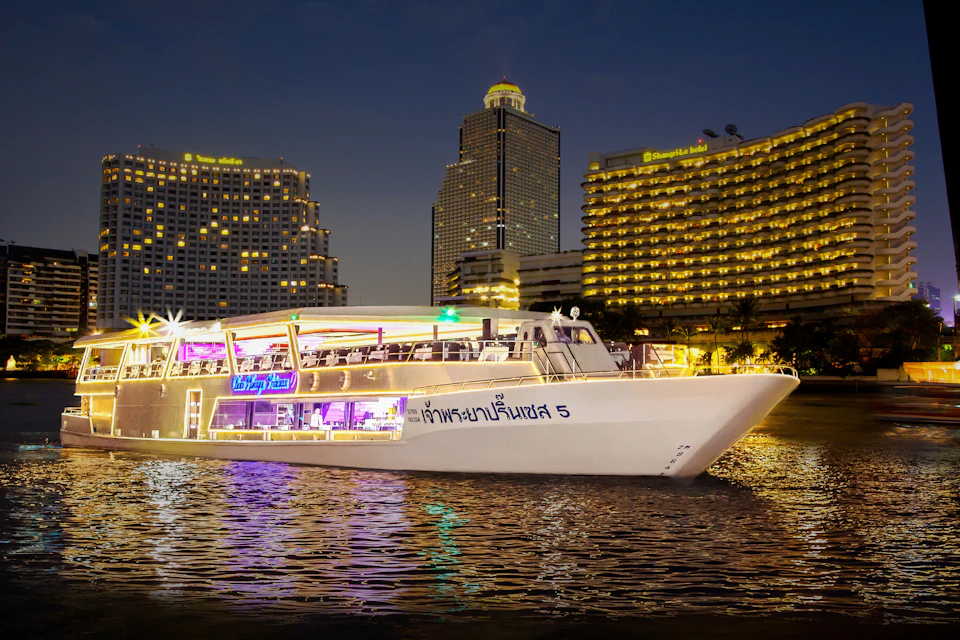 A brightly lit dinner cruise boat on water at night, with illuminated modern buildings in the background.